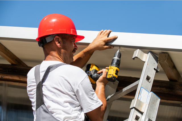 Roof repair worker fixing side of the roof on a ladder with a drill bit.